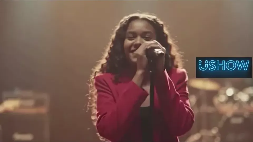 Uma estudante canta no palco de um festival escolar, sob a luz dos refletores.